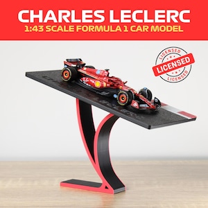 Könnte beinhalten: Ein rotes und schwarzes Modell eines Formel-1-Rennwagens im Maßstab 1:43, montiert auf einem schwarz-roten Ständer. Das Modellauto hat Sponsorenlogos und die Aufschrift "CHARLES LECLERC". Der Ständer hat ein weiß-rotes Streifendetail.