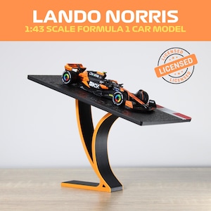 Könnte beinhalten: Ein Modellauto im Maßstab 1:43 eines Formel-1-Rennwagens in Schwarz und Orange, präsentiert auf einem schwarz-orangenen Ständer. Der Text "Lando Norris" und "Licensed" sind im Bild zu sehen.