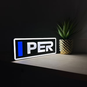 May include: A black rectangular sign with white lettering that reads "I PER" and a blue vertical bar to the left of the text. The sign is illuminated from behind, creating a bright glow. The sign is sitting on a light wood surface.