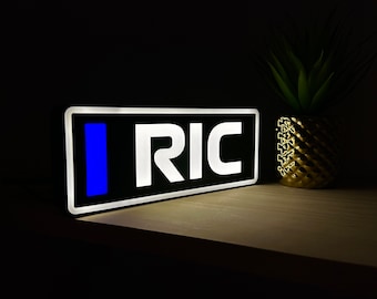 May include: A black light-up sign with white letters spelling "RIC" and a blue vertical bar to the left of the letters.