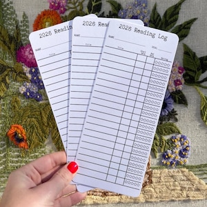 May include: Three white 2026 Reading Log cards are held in a hand with red nail polish. Each card has a section for name, title, and goal, with a rating section. The background features an embroidered floral design with pink, purple, and orange flowers.