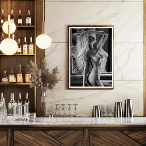 May include: Black and white photograph of a woman standing in front of a mirror, with her back to the camera. She is wearing a white shirt and has long, flowing hair. The photograph is framed in a black frame.
