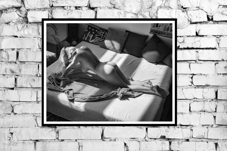 以下が含まれることがあります： 縞模様の毛布を敷いたベッドに横たわる人の白黒写真。その人は裸で、カメラに背を向けている。ベッドは白いシーツと枕で整えられている。ベッドにはいくつかの枕があり、その中には赤、白、青のパターンがあるものもある。