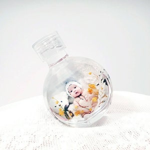 以下が含まれることがあります： 赤ちゃんとおもちゃのテディベアが入った透明なガラス瓶。赤ちゃんは白い帽子と明るい色の服を着ています。瓶の中には黄色い花やその他の装飾要素もあります。