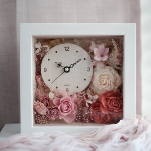 May include: A square white framed clock with a light wood-colored clock face. The clock face has black hands and the word "QUARTZ" printed below the center. The clock is filled with pink and red roses and other dried flowers.