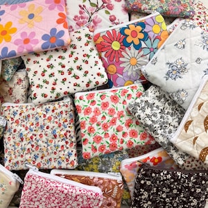 May include: A selection of small, quilted fabric pouches with diverse floral patterns. The pouches display a range of colours, including pink, red, blue, and yellow, with white zips. The designs include daisies, cherry blossoms, and other stylised flowers.