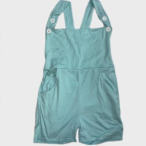 May include: A light turquoise romper featuring a square neckline, wide shoulder straps, and two white buttons on each strap. The garment includes two side pockets and a short-length design, providing a simple and functional one-piece outfit.