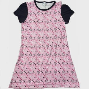 May include: A pink nightgown with a repeating pattern of cartoon cat faces, hearts, and dots. The dress has short black sleeves and a black trim around the neckline. The overall design is playful and cute.