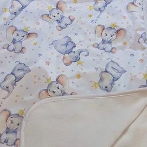 Può includere: Una copertina per neonati bianca con una stampa di elefanti con corone e stelle in blu chiaro e giallo. La copertina ha una texture morbida e soffice.
