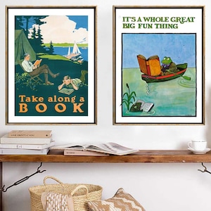 May include: Two framed wall art prints featuring vintage-style illustrations. The left print shows people reading outdoors with the text Take along a BOOK. The right print depicts a frog in a boat reading with the text IT'S A WHOLE GREAT BIG FUN THING.