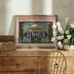 K&ouml;nnte beinhalten: Gerahmtes Vintage-Poster f&uuml;r die Produktion von "Ben-Hur" im Illinois Theatre, Chicago, vom 2. September. Das Poster, in einem goldenen Rahmen, zeigt eine Schiffsillustration. Es steht auf einer Holzkommode mit Korb und Blumen.