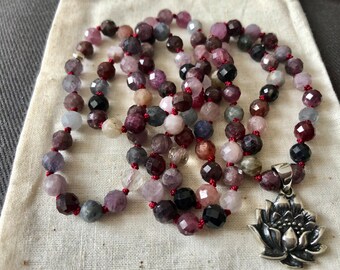 May include: A multi-colored gemstone mala necklace with a silver lotus flower pendant. The necklace is made with red, pink, purple, and blue gemstones.