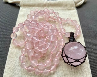 May include: A pink rose quartz mala necklace with a large rose quartz pendant wrapped in brown wire. The necklace is made of round beads and is lying on a white fabric surface.