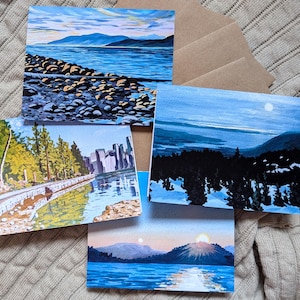 May include: Four watercolour landscape paintings depicting tranquil natural scenes. The paintings show a coastal view, a city skyline reflected in water, a snowy mountain range, and a sunset over a lake. Brown envelopes are visible.