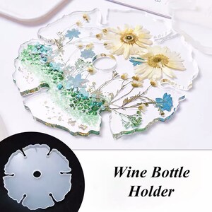 May include: A clear resin wine bottle holder with a floral design. The holder features dried flowers and leaves in shades of white, blue, and green. The holder is shaped like a leaf and has a hole in the center for the bottle neck. The text "Wine Bottle Holder" is printed below the holder.