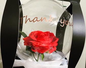Peut inclure: Une bulle en plastique transparente contenant une seule rose rouge. La bulle est entourée de papier de soie blanc et porte l'inscription "Merci" en lettres dorées.