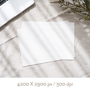 Puede incluir: Dos hojas blancas en blanco sobre un fondo de tela de lino beige claro. El papel est&aacute; colocado plano sobre la tela con un ordenador port&aacute;til en la esquina superior izquierda de la imagen. El port&aacute;til est&aacute; cerrado y la pantalla est&aacute; orientada hacia arriba. La tela tiene una textura natural y est&aacute; iluminada por la luz del sol, creando una luz suave y natural.
