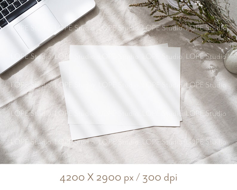 Puede incluir: Dos hojas blancas en blanco sobre un fondo de tela de lino beige claro. El papel est&aacute; colocado plano sobre la tela con un ordenador port&aacute;til en la esquina superior izquierda de la imagen. El port&aacute;til est&aacute; cerrado y la pantalla est&aacute; orientada hacia arriba. La tela tiene una textura natural y est&aacute; iluminada por la luz del sol, creando una luz suave y natural.