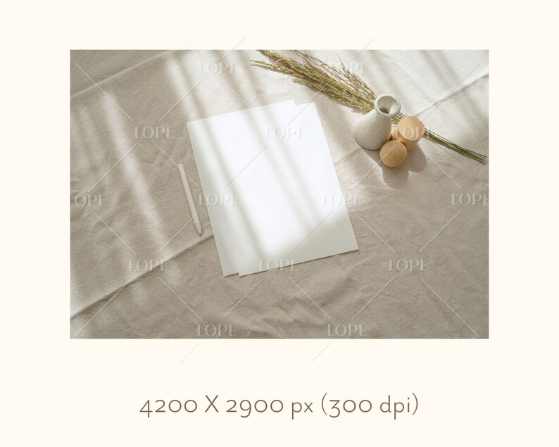 May include: A white sheet of paper with a white stylus on a beige linen fabric background. A small white ceramic vase with dried grass and three wooden spheres are in the top right corner of the image.