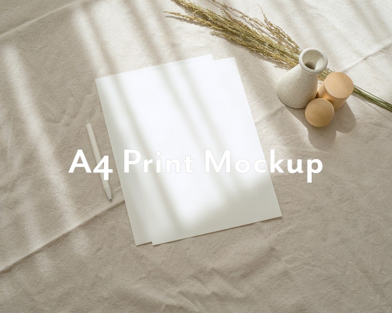 May include: A white A4 sheet of paper on a beige fabric surface with a white pen and a small vase with dried flowers. The text "A4 Print Mockup" is printed on the paper.