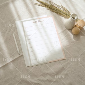 May include: A weekly planner with a pink and white cover, a white pen, and two wooden decorative objects on a beige fabric surface.