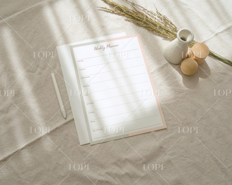 May include: A weekly planner with a pink and white cover, a white pen, and two wooden decorative objects on a beige fabric surface.