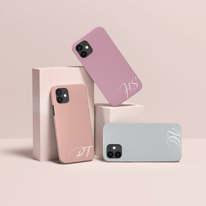 Puede incluir: Tres fundas para teléfono con diferentes iniciales, "HS", "RT" y "DV", en escritura blanca. Las fundas son rosa, melocotón y gris claro.