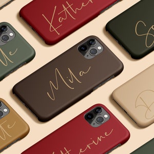 Puede incluir: Una colección de fundas personalizadas para teléfonos en varios colores, incluyendo verde, marrón y rojo. Cada funda presenta un nombre en una fuente cursiva elegante. Las fundas están diseñadas para un smartphone y tienen un recorte para la cámara.