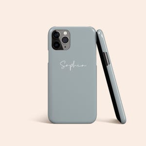 Puede incluir: Una funda de teléfono azul claro con el nombre "Sophia" en cursiva. La funda está apoyada contra otra, revelando el interior negro. La funda del teléfono tiene un módulo de cámara.