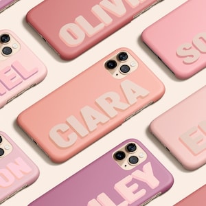 Puede incluir: Una colección de fundas de teléfono rosa y morado con diferentes nombres impresos en una fuente en negrita en 3D. Las fundas están dispuestas en una cuadrícula sobre un fondo beige claro.