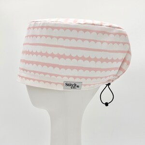 May include: Pink and white striped scrub hat with a scalloped pattern. The hat has a drawstring and a small label that says "Stitch & Co."