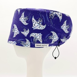 May include: Purple scrub hat with a white and blue patterned design of manta rays. The hat has a drawstring and a label that says "Stitch and co."