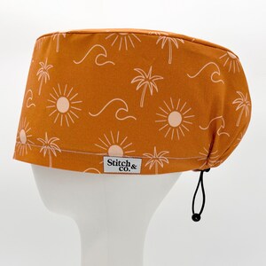 May include: A brown scrub cap featuring a repeating pattern of suns, palm trees, and waves in a light beige hue. The cap has a white label reading "Stitch & co." and a black drawstring for adjustment.