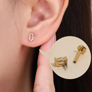 May include: A gold-toned stud earring featuring an oval-shaped clear gemstone. The earring is shown inserted into an earlobe, with an inset circular detail displaying the side profile of the stud and its flat-backed post mechanism.