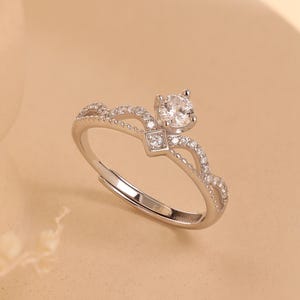 May include: A silver-toned ring with a crown design, featuring a large, clear gemstone at the top and smaller stones along the band. The ring is set against a neutral background, showcasing its delicate details. The ring is an adjustable size.
