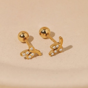 May include: A pair of gold-colored stud earrings. Each earring features a small, leaf-like design with embedded clear crystals. The earrings have a ball post and are displayed on a light beige surface.