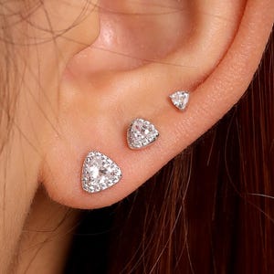 Puede incluir: Primer plano de una oreja con tres pendientes de plata. Los pendientes tienen forma de triángulo y están incrustados con pequeños cristales transparentes. La oreja está sobre un fondo borroso.