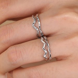 May include: Two silver rings with wave and zigzag designs are worn on a person's finger. The rings are delicate and have a shiny finish. The person is holding a white teacup and saucer.