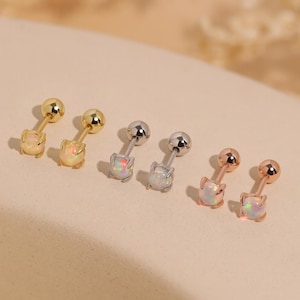 May include: A collection of six small stud earrings. Two are gold-toned, two are silver-toned, and two are rose gold-toned. Each earring features a small, round ball and a cat-shaped opal stone. The earrings are displayed on a neutral-toned surface.