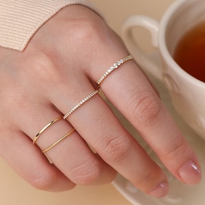 May include: A hand wearing four gold rings, including a band with small clear stones. A white teacup filled with tea sits in the background. The rings are delicate and elegant, suggesting a jewelry theme.