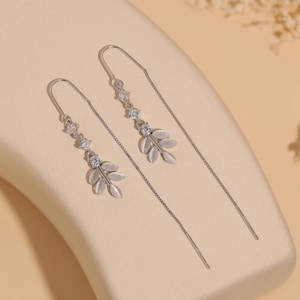 Pode incluir: Um par de brincos de fio de prata. Cada brinco apresenta um design delicado com três pedras preciosas redondas e transparentes e um grupo de três pingentes em forma de folha. Os brincos são exibidos em uma superfície bege.