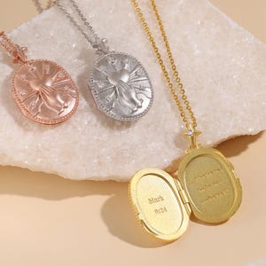 Puede incluir: Tres collares con relicario ovalado en tonos oro rosa, plata y oro. Cada uno presenta un diseño en relieve de manos en oración y un corazón con rayos de estrella. El relicario dorado está abierto, revelando el texto grabado Mark 9:24 y un pasaje.