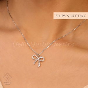 May include: A silver necklace with a bow pendant, adorned with clear crystals. The delicate chain and bow design create an elegant look. The text "SHIPS NEXT DAY" is visible in the image.