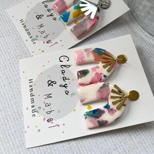 Peut inclure: Une paire de boucles d'oreilles faites à la main avec un motif d'arche marbré blanc, rose et bleu avec des accents dorés. Les boucles d'oreilles sont sur une carte blanche avec le texte "Lady & Mabel Handmade".