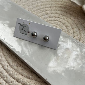 May include: Two small, silver-coloured ball stud earrings. The earrings are presented on a white card with the text "Clodya & Mabel Handmade". The card rests on a light grey rectangular surface.