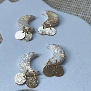Peut inclure: Une paire de boucles d'oreilles en forme de croissant de lune dorées et blanches avec des cercles dorés pendants. Les boucles d'oreilles sont en pâte polymère et ont un effet marbré.
