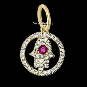 Puede incluir: Colgante dorado con un diseño de mano de Hamsa, rodeado por un anillo de pequeñas piedras preciosas transparentes. La mano de Hamsa tiene una piedra preciosa roja redonda en el centro. El colgante tiene un lazo para una cadena. El texto "Santoshdesign" es visible.
