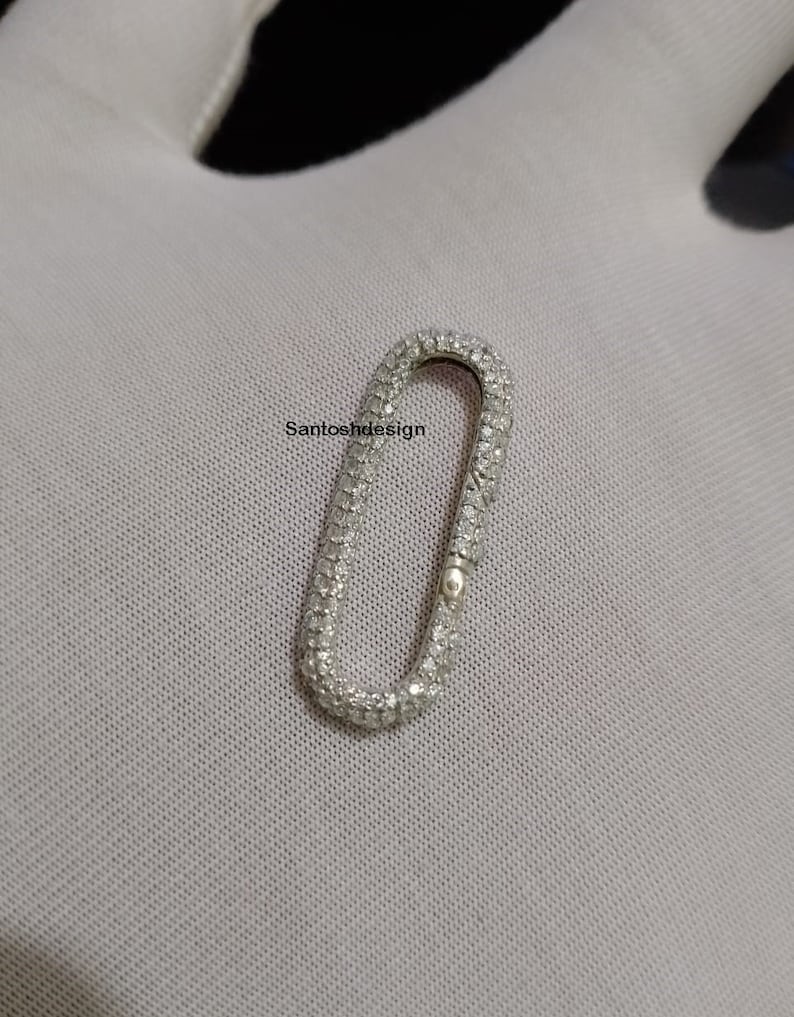 May include: A silver oval-shaped charm with a small loop at the top. The charm is covered in small, clear rhinestones.