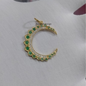 Pode incluir: Um pingente de lua crescente de ouro com pedras preciosas verdes e pedras de zircônia cúbica brancas. O pingente está sobre um fundo branco. Santoshdesign está escrito no canto superior direito da imagem.