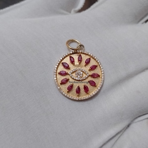 May include: Gold pendant with a round design featuring a stylized eye surrounded by red gemstones and a halo of smaller white gemstones.